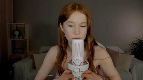 Media: Video of a red-haired girl in a pink dress, singing into a white microphone, in a dimly lit living room with a gray couch and a bookshelf.