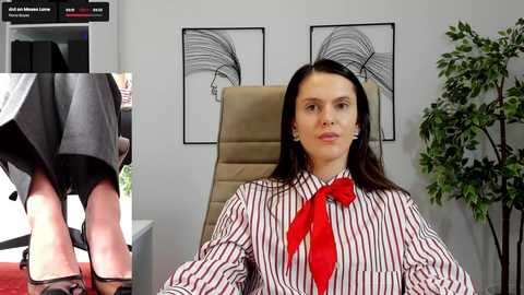 Media: Video of a woman with long brown hair, wearing a red-striped blouse, red tie, and black skirt, seated in an office with beige chair, black-and-white abstract art on white walls.