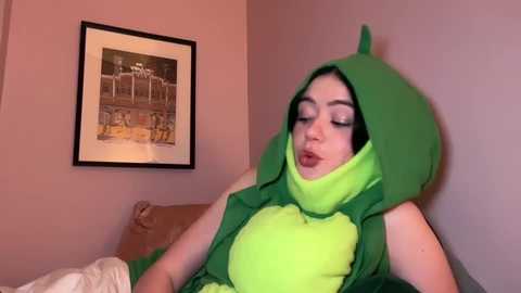 Media: Video of a young woman in a green, plush dinosaur costume with a hood, lying on a bed with a beige pillow and framed artwork on the wall.