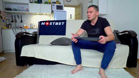 Media: Video of a fit, light-skinned man with short brown hair, wearing a black t-shirt and blue jeans, playing a video game on a couch in a modern, minimalist living room.