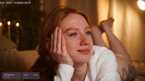 Media: Video of a red-haired woman with fair skin, lying on a bed, resting her head on her hand, in a cozy, warmly-lit room with a soft focus.