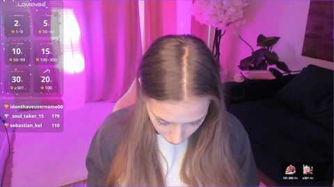 Media: Video of a young woman with straight, light brown hair, wearing a gray top, looking downward, surrounded by a pink-lit bedroom with a floral arrangement and a bed in the background.