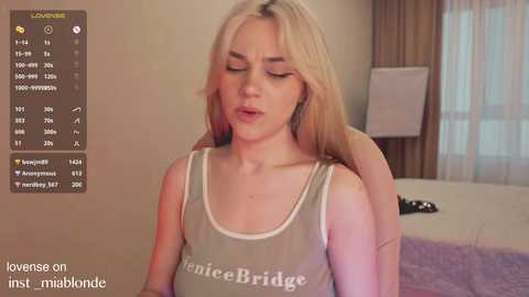 Media: Video of a blonde woman with long hair wearing a \"Francesca Bridge\" tank top, seated on a bed, with a man's hand on her shoulder. Background shows a bedroom with a white bed, beige walls, and a window.