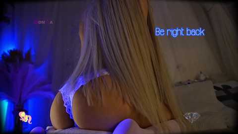 Media: Video of a blonde woman in white lace lingerie, lying on a bed with dim blue lighting, caption reading \"Be right back.\