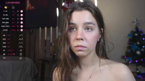 Media: Video of a young woman with long brown hair, light skin, and freckles, wearing a necklace. Background features a dimly lit room with a TV screen displaying live streaming statistics.