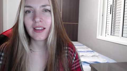 Media: Video of a young Caucasian woman with long, straight, blonde hair, wearing a red plaid shirt, sitting on a bed with white sheets and a window with blinds in a modern bedroom.