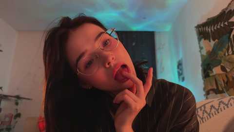 Media: Video of a young woman with long black hair, fair skin, and round glasses, licking her finger, in a dimly lit room with greenish-blue LED lights, and a messy background.