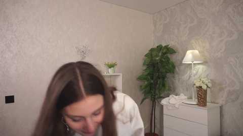 Media: Video of a woman with long brown hair, wearing a white dress, in a room with floral wallpaper, a green plant, and a white lamp on a dresser.