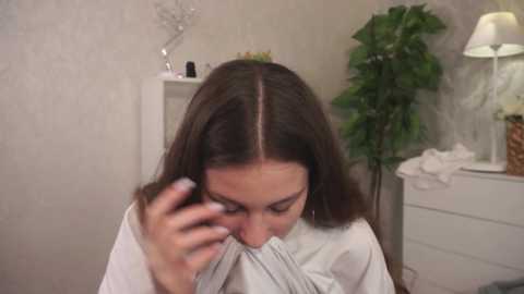 Media: Video of a woman with shoulder-length brown hair, wearing a white robe, holding a phone to her ear, standing in a beige-tiled bathroom with a white dresser and green plant in the background.