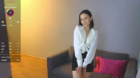 Media: Video of a smiling Asian woman in a white blouse, black shorts, standing in a modern living room with gray sofa, wooden furniture, and colorful pillows.