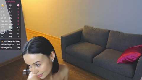 Media: Video of a young woman with fair skin, dark hair, and no top, sitting on a wooden floor near a grey couch with a red pillow. A smartphone screen showing social media accounts is visible in the background.