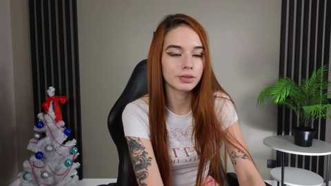 Media: A video of a fair-skinned woman with long red hair, wearing a white T-shirt, sitting on a black gaming chair in a room with a Christmas tree, potted plants, and dark curtains.