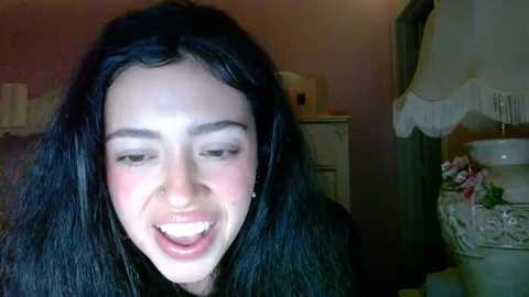 Media: A candid video captures a young woman with long black hair, fair skin, and a nose ring, laughing in a dimly lit bathroom with a white sink, floral decor, and a mirror.