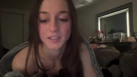 Media: Video of a young woman with long brown hair, wearing a gray jacket, indoors. She has fair skin and a soft smile. Background includes a mirror, nightstand, and dim lighting.