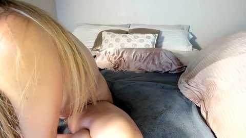 Media: Video of a blonde woman, topless, lying on a bed with gray bedding, pillows, and a beige blanket, in a dimly lit room with a white wall.