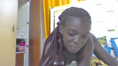 Media: A video of a dark-skinned woman with braided hair, wearing a sheer brown top, leaning on her hand, in a cluttered room with yellow curtains, shelves, and colorful bags in the background.