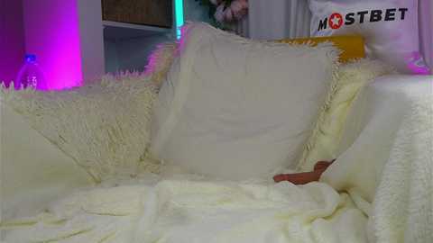 Media: Video of a plush, white, furry sofa with a cream-colored pillow, purple and blue ambient lighting, and a \"MOSTBET\" sign in the background.