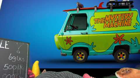 Media: Video of a bright, colorful Mystery Machine van with a blue and green exterior, featuring a cartoonish design. The background is a vivid blue, with a black chalkboard partially visible in the foreground, displaying \"BOO\" and \"SUCK.\