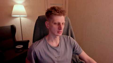 Media: Video of a fair-skinned, young man with spiky, light brown hair, wearing a gray T-shirt, sitting in a striped armchair in a dimly lit room with a lamp and beige walls.