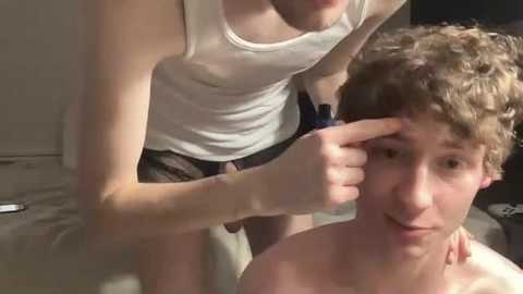 Media: Video of two young men, one with curly hair, shirtless, and the other in a white tank top, grooming the first's hair in a bedroom.