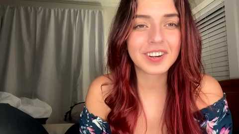 Media: Video of a young woman with long, wavy, red-brown hair, fair skin, and a warm smile. She wears a dark floral off-shoulder top. Background includes white curtains, a bed with white sheets, and a window with closed blinds.
