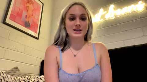 Media: Video of a young Caucasian woman with platinum blonde hair, wearing a light blue lace bralette, standing in a cozy room with a brick wall, a framed painting, and string lights.