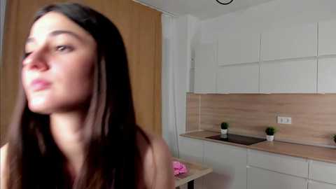 Media: Video of a young woman with long brown hair, wearing a pink shirt, standing in a modern kitchen with light wood cabinets and white countertops.