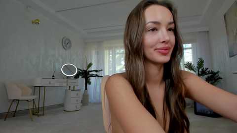 Media: Video of a smiling, fair-skinned woman with long brown hair, wearing a sleeveless top, in a bright, modern room with white walls, a round mirror, and plants.