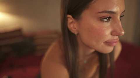 Media: Video of a young, light-skinned woman with straight, dark brown hair, wearing a red top, gazing pensively. Background shows a blurred, warmly lit room with a bed and wooden furniture.