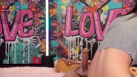 Media: Video of a person with light skin, wearing a gray shirt, holding a realistic, erect, brown dildo, against a backdrop of vibrant, graffiti-style \"LOVE\" letters.