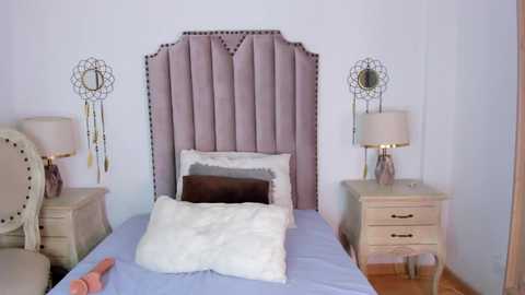 Media: Video of a modern, minimalist bedroom with a tufted, light purple headboard, beige upholstered chairs, white bedding, and two matching nightstands with gold lampshades and dreamcatcher wall decorations.