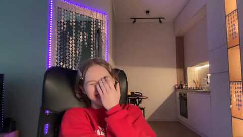 Media: Video of a young girl with light skin and brown hair, wearing a red sweater, laughing with her hand over her mouth in a modern, dimly lit living room with a black leather chair, purple LED strip lights, and a kitchenette.