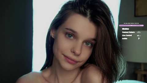 Media: Video of a young woman with fair skin and long brown hair, wearing a subtle smile. She has blue eyes and is topless, with a blurred background featuring a lamp and a white wall.