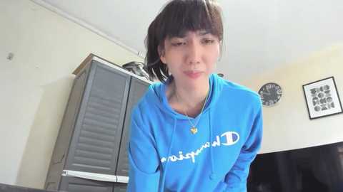 A video of an Asian woman with short brown hair, wearing a blue Adidas hoodie, standing in a room with a gray refrigerator, black clock, and framed art.