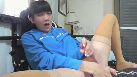 Video of an Asian woman with long black hair, wearing a blue sweatshirt, beige thigh-high stockings, and a shocked expression, masturbating on a bed in a dimly lit room.