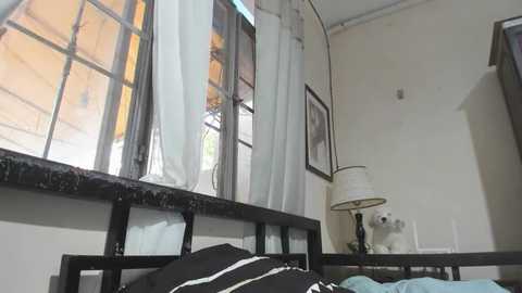 Media: A video of a cozy bedroom with a black metal bed, a white lamp, and a window with sheer curtains. The room is painted white with a vintage feel.