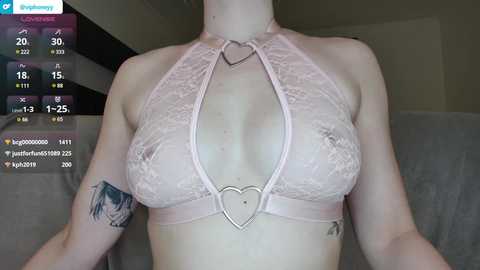 Media: Video of a fair-skinned woman with tattoos, wearing a pink lace bra with a heart-shaped cutout, and a silver heart pendant necklace. Background shows a bathroom with a shower curtain.