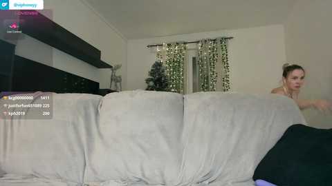 Media: Video of a woman with light skin, brown hair, and a slender build sitting on a gray sofa in a living room. The room has a minimalist decor with white walls, a Christmas tree, and a black shelf.