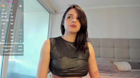 Media: Video of a young woman with straight black hair, wearing a black sleeveless top and white skirt, standing in a modern bedroom with a grey upholstered headboard and a bed.