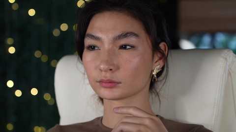 Media: Video of a young Asian woman with straight black hair, wearing gold butterfly earrings, sitting on a white chair with a blurred green and gold bokeh background.