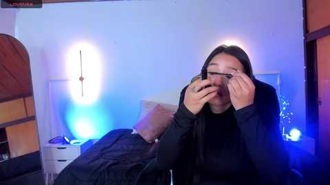 Media: A video shows a young woman with long dark hair, wearing glasses and a black top, adjusting her glasses in a dimly lit bedroom with blue and white ambient lighting.