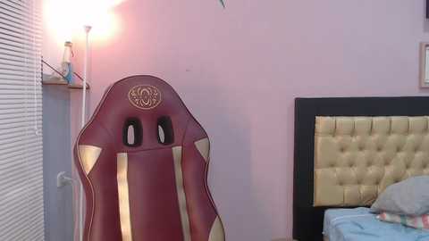 Media: Video of a maroon gaming chair with gold accents, placed in a pink-walled room with a beige tufted headboard, beside a bed with light blue bedding.