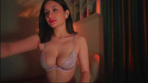 Media: Video of a young Asian woman with straight black hair, wearing a light purple bra with sequins, standing indoors with a dim, warm light, and blurred background.