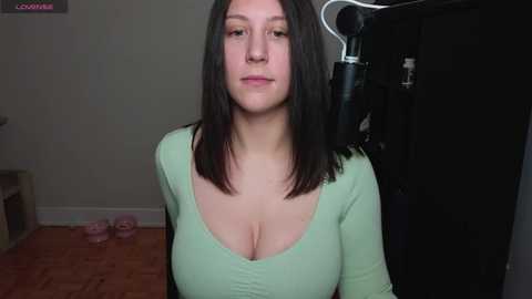 Media: A video of a young woman with medium-length dark hair, wearing a low-cut green top, sitting in a room with a black exercise bike.