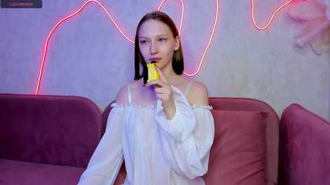 Media: Video of a slender young woman with straight, shoulder-length brown hair, wearing a white off-shoulder blouse, eating a banana, seated on a maroon couch, in a dimly lit room with neon pink lighting and a textured wall.