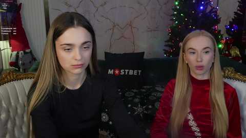 Media: Video of two young women, one in a black sweater, the other in a red velvet top, sitting on a sofa with a decorated Christmas tree and gifts in the background.