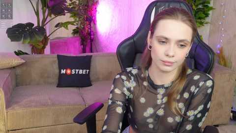 Media: A video of a young, pale-skinned woman with light brown hair, wearing a sheer floral top, sitting in a black gaming chair with a purple light background.