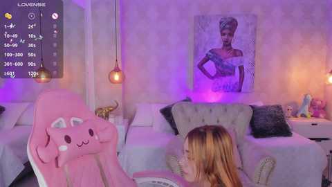 Media: Video of a young woman with blonde hair sitting on a pink plush chair in a cozy, purple-lit bedroom with a framed portrait and a bed with white linens and pillows.