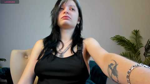 Media: Video of a fair-skinned woman with long black hair, wearing a black tank top, silver necklace, and bracelet, with a tattoo on her left arm. Background features a beige chair, green plant, and neutral walls.