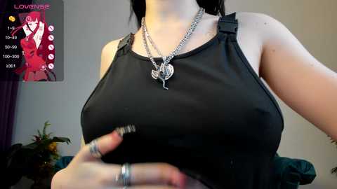 Media: Video of a fair-skinned woman with large breasts in a black, sleeveless top. She wears a silver necklace with a pendant. Background shows a pink poster and a green couch.